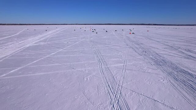 Aerial flying view of the frozen Simcoe lake showing the snowmobile tracks and ice fishing huts village in the snow. Ice fishing huts or cabins in scenic aerial shot. Charter shelters for ice fishers