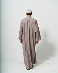 A man is seen from behind walking away while wearing a traditional long robe. He is also wearing a white cap and appears to be in a brightly lit, minimalist setting.