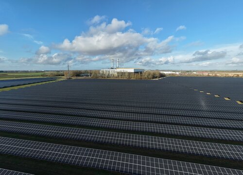 Aerial view of gleaming solar panels stretch across the landscape towards a distant industrial building under a sky dotted with fluffy clouds, Sutton Bridge, Sutton Bridge, United Kingdom.