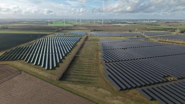 Aerial view of vast solar panels gleam, reflecting the sky, contrasting with the earth tones and distant wind turbines, Sutton Bridge, Sutton Bridge, United Kingdom.
