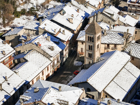 Aerial view of snow-dusted rooftops and the striking bell tower of a church, a red car contrasting against the white and grey, Exilles, Piemonte, Italy.