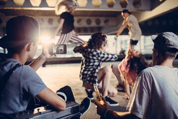 Young friends filming street dancers under urban bridge