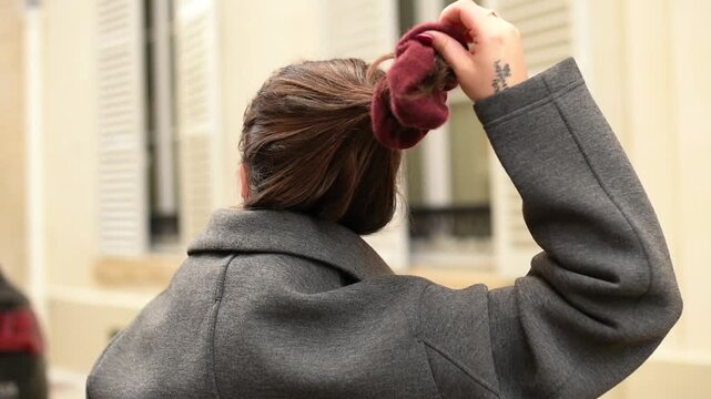 Back view of a woman with brown hair removing a burgundy scrunchie from her ponytail while wearing a grey coat outdoors.