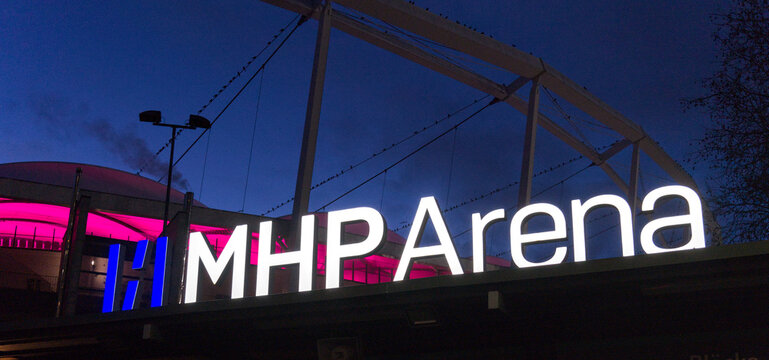 The glowing 'MHP Arena' lettering shines brightly against the illuminated facade of the stadium building on a dark night.