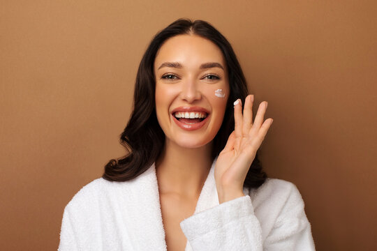 Beauty routine. Happy woman applying facial cream on her cheeks with joyful expression, doing daily skincare, standing against beige background