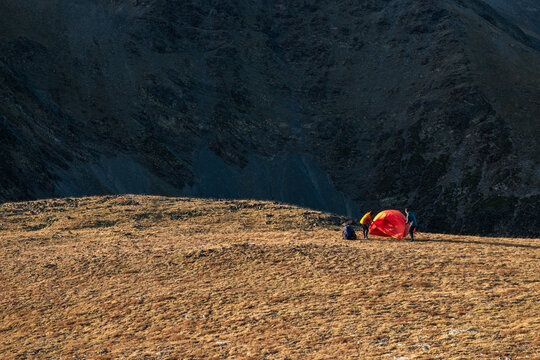 Hiking campers pitching red tent within vast alpine mountain bowl