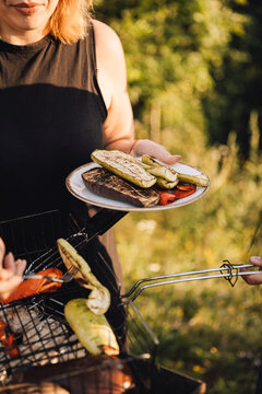 Woman serving grilled vegetables from barbecue, holding plate outdoors in summer sunlight