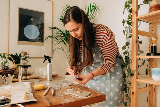 Focused woman sculpting clay fish with tools at wooden workbench