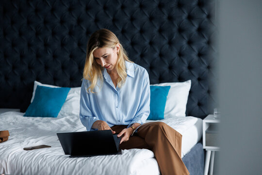 Pretty concentrated adult businesswoman using laptop sitting on bed