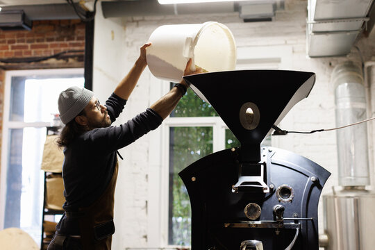 Male worker pouring coffee beans from bucket in roaster machine