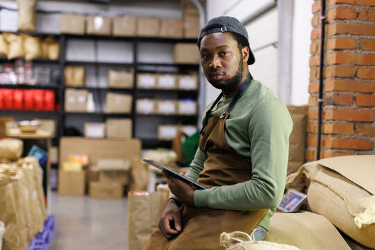 Male worker with tablet on sacks with coffee and looking at camera