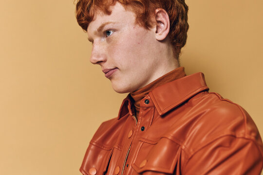 Portrait of young man with red hair wearing brown leather jacket against beige background. Profile view, casual fashion, natural expression, studio lighting, closeup headshot.