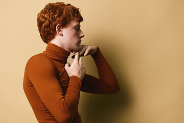 Young redhead person adjusting turtleneck sweater against beige background, side profile portrait with casual style and minimalist warm color tones. © SHOTPRIME STUDIO