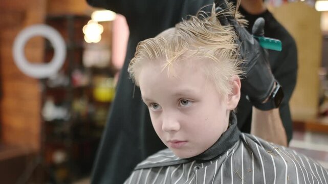 Cute school age boy getting haircut in barbershop. Children hairdresser with professional tools - comb and scissors. Cutting hair for kids. Services of a professional stylist.