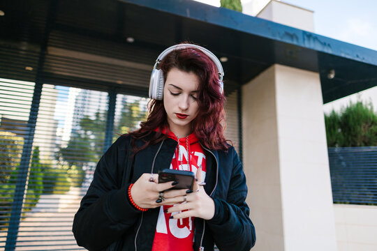 Young woman wearing headphones scrolling smartphone screen outdoors