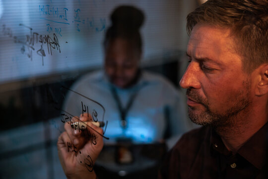 Man writes equations on glass, colleague works in background.