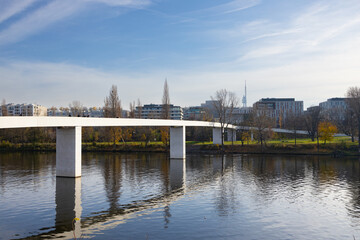 Obraz premium Modern pedestrian bridge spanning a calm river with reflections, surrounded by urban buildings and trees under a clear blue sky in a city landscape
