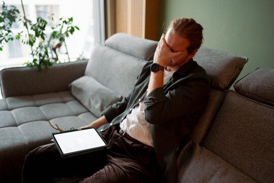 Man sitting on couch covers face with hand in modern living room
