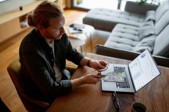 Man counts cash while working on laptop at home office
