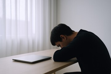 Naklejka premium Young adult experiencing burnout, head resting on desk with laptop in simple, bright interior