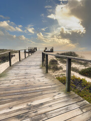 Obraz premium A wooden boardwalk extends in perspective over sandy dunes and coastal vegetation towards a distant viewing platform at Furadouro beach Ovar Portugal under a bright, dramatic, cloudy sky