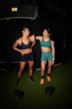 Two women smiling and talking in a gym setting during workout ti