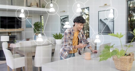 Holding phone, man wearing plaid shirt, yellow tee tapping laptop in kitchen with network icons