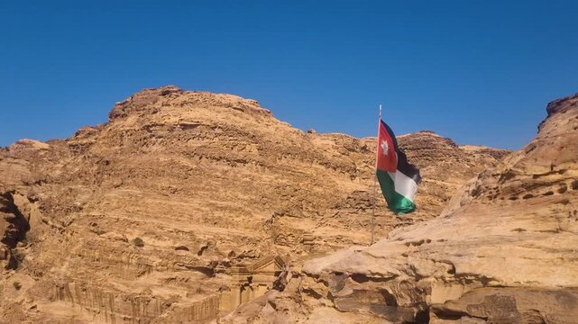 A Jordanian flag waves in the wind against desert rock formations near Wadi Musa, under a clear blue sky, symbolizing national pride and the iconic landscapes of Jordan