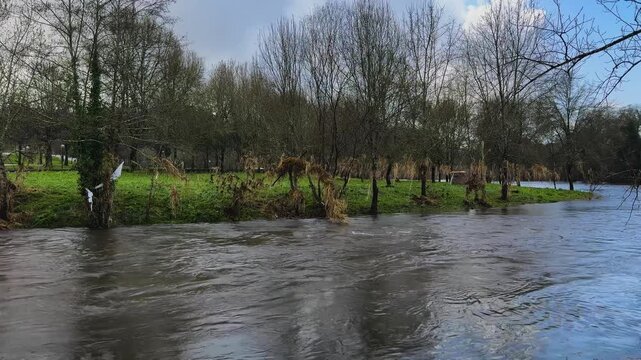 Rushing water flowing through a normally calm river, with debris in the current as the water level rises dangerously high.