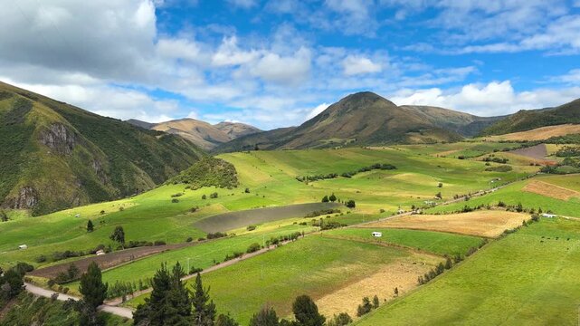 Handheld wide shot of a dirt road winding through green agricultural fields in a sunny Andean valley with mountains and blue sky in Ecuador.