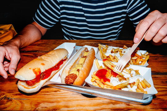 a man eats french fries from a paper container next to hot dogs