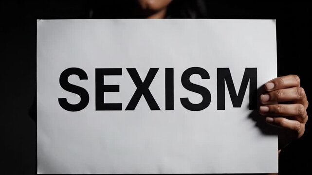 Minimalist cinematic video of a hand presenting a white paper sign with the word SEXISM in bold black letters identity hidden dark background dramatic lighting emphasizing message smooth slow camera m