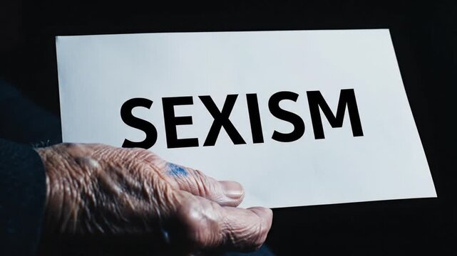 Dramatic cinematic video emphasizing social awareness showing a hand holding a white paper with bold black SEXISM text dark moody background soft shadows smooth slow camera movement medium close up sh
