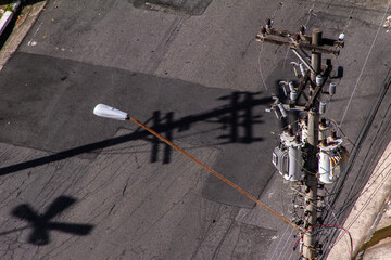 High-angle view of a utility pole with transformers and a street lamp, casting strong shadows on the asphalt. A concept of urban infrastructure, power distribution, and city textures.