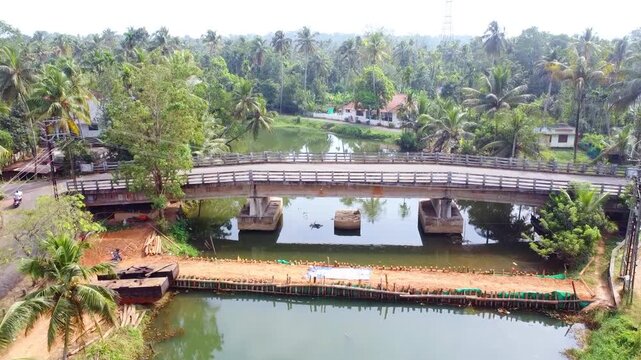 This aerial footage captures a serene river bridge nestled within a lush, tropical landscape. It features a modern concrete bridge, a temporary construction bund, and vibrant coconut groves.