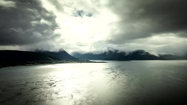 Cinematic drone hyperlapse over shimmering sea and moody clouds near Sortland, Vester&aring;len, Northern Norway, with dramatic light breaking across water and distant mountains