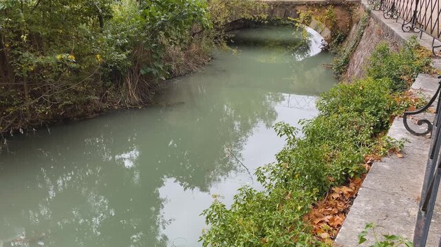 Images of a branch of the Tagus River that borders the Royal Palace of Aranjuez.The river flows with whitish waters,with riverside vegetation on one side and a wall with a wrought-iron railing