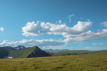 Obraz premium Sunlit grassy meadow near abyss edge and wavy green hills far away in sunny day. Layered undulating hilly relief in sunny day. Pointy peak under puffy clouds in blue sky. Big peaked top in bright sun.
