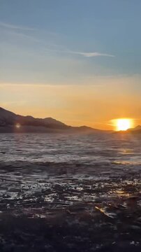 A cinematic vertical handheld shot of a breathtaking sunrise over the mountains at Sayram Lake, Xinjiang, China. The golden sun is rising above the horizon, casting a warm glow on the crystal-clear bl