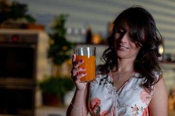 Woman holds glowing beverage joyfully, Caucasian female enjoying bright juice in kitchen, Amber liquid radiates warmth while woman gazes with subtle happiness in background