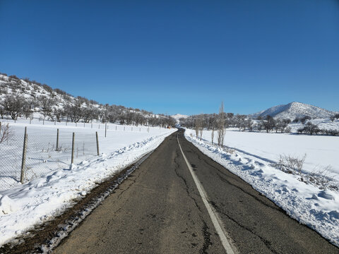 Straight winter road stretching through snowy fields with distant mountains and clear blue sky, capturing the calm beauty and bright stillness of a cold winter day.  
📍Marivan, Kurdistan 