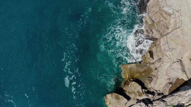 Top Down Aerial View of Waves Crashing on Rocky Coastline with Turquoise Sea Water. Drone Shot Highlighting Coast Erosion caused by constant sea wave impact.