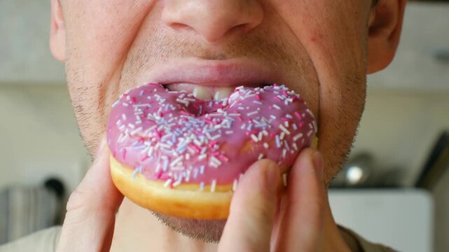 Close up man eating delicious sweet donut with pink icing, colorful sprinkles, taking large bite, chewing sugary junk food with great pleasure in mouth. Man mouth eating unhealthy pink glazed donut