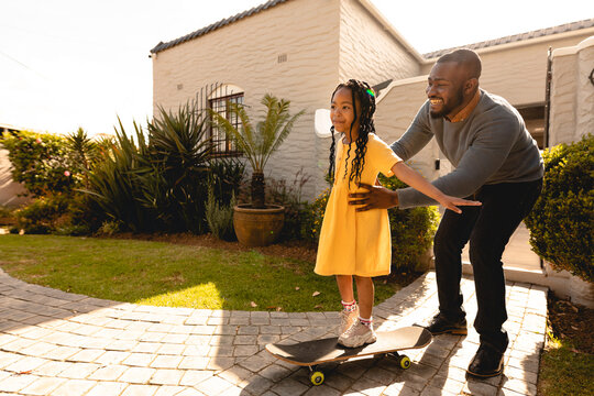 Cheerful african american man supporting daughter learning skateboard in backyard during weekend