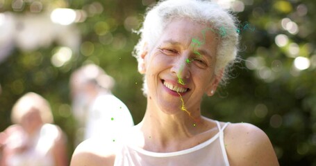 Smiling senior woman wearing white top showing paint specks across face in garden, pearls and bokeh