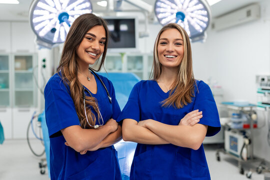 Two beautiful and confident young female medical professionals in blue scrubs stand with arms crossed in a modern surgical theater. Team of friendly nurses and doctors providing healthcare.