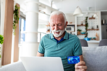 Fototapeta premium Senior Man Making Online Payment with Credit Card. E-commerce and Financial Technology, Retirement Lifestyle concept. Happy elderly male using laptop and bank card for shopping while on sofa.