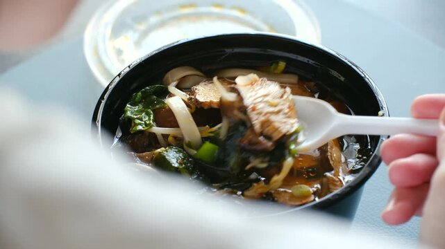 Close up hands stirring, eating traditional japanese miso soup with shiitake mushrooms, tofu, noodles from black takeout bowl with plastic white spoon. Person eating hot japanese miso soup for lunch