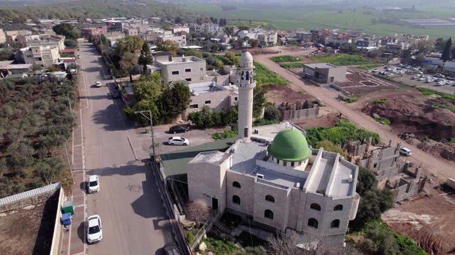 Aerial drone shot of a mosque in the Wadi Ara region of northern Israel, featuring a green dome and tall minaret as pigeons fly around the structure, with surrounding village buildings, roads, and agr