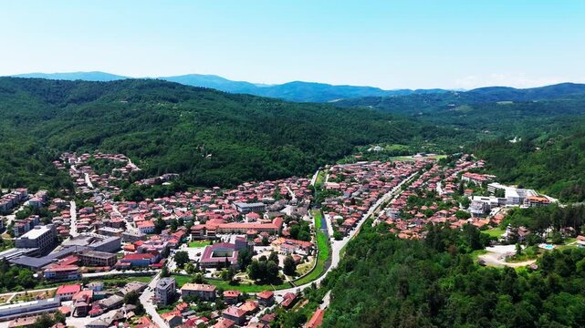 Drone curves along Tryavna&rsquo;s layout, showcasing terracotta roofs, parks, river, and surrounding green hills shaping the village valley.